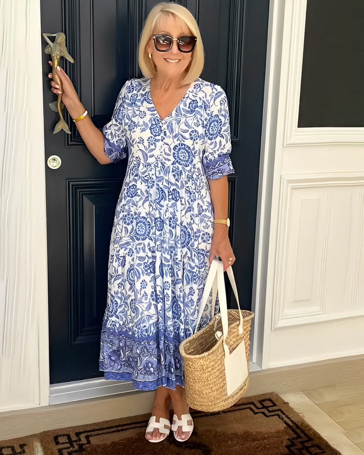 Woman in a blue and white dress holding a straw bag, standing in front of a dark door.