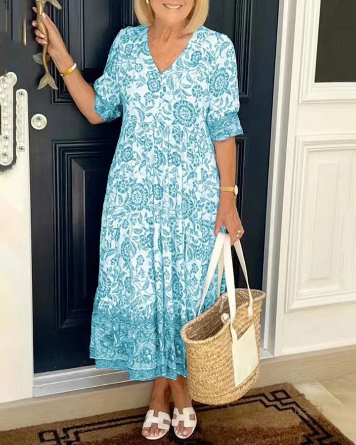 Woman in a blue floral dress holding a straw bag, standing in front of a dark door.