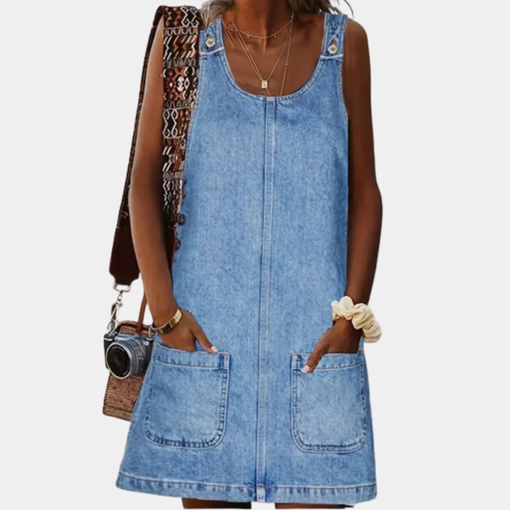 Person wearing a denim dress holding a camera and a patterned bag on a white background