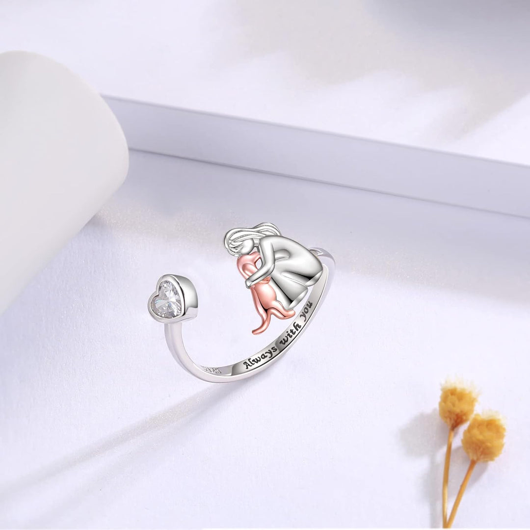 Silver ring with a heart and pink figure on a white surface with dried flowers.