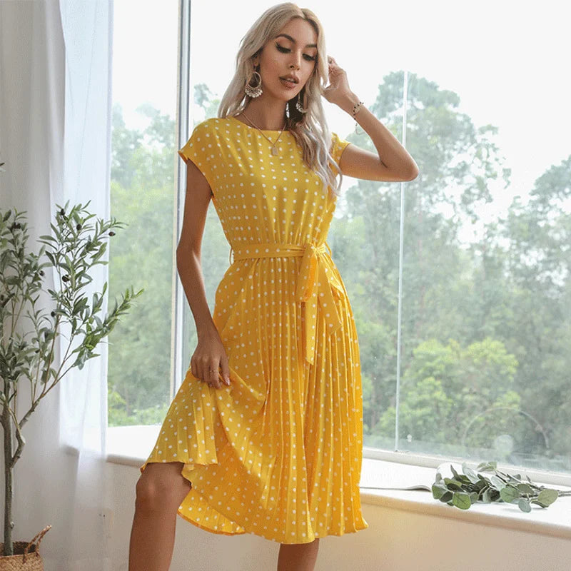 Woman wearing a yellow polka dot dress standing by a window with greenery outside.