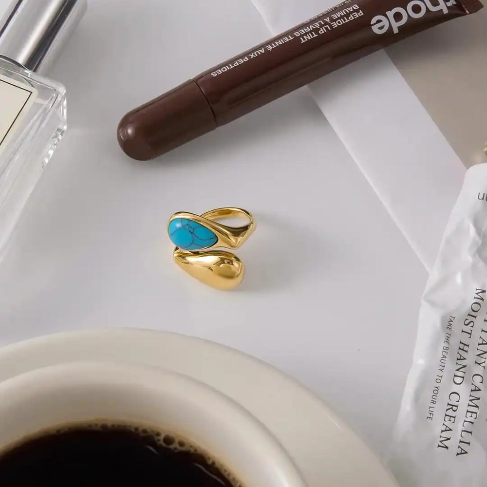 Gold earrings with a blue gemstone on a white surface with a cup of coffee, pen, and hand cream.