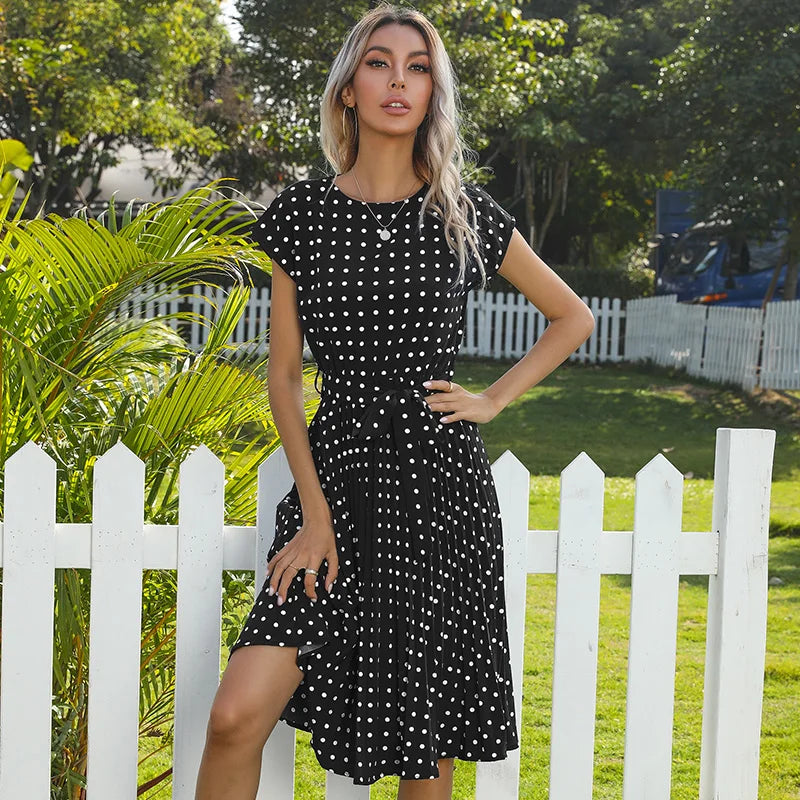 Woman in a black polka dot dress standing in front of a white picket fence with greenery.