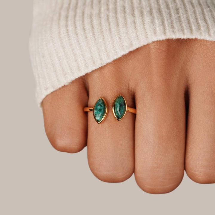 Gold ring with green gemstones on a finger against a neutral background