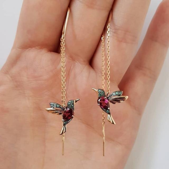Gold hummingbird earrings with gemstones on a hand against a light background
