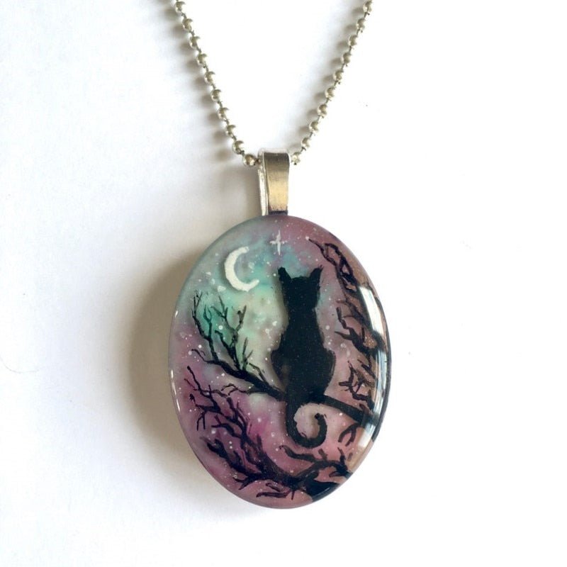 Oval-shaped pendant with a black cat silhouette, tree, and moon design on a white background.
